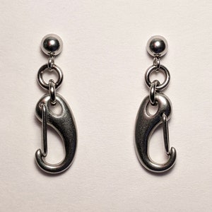 Peut inclure: Une paire de boucles d'oreilles argentées avec une petite boule en haut et un charme en forme de crochet suspendu en dessous.