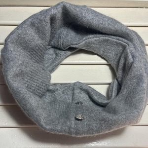 Puede incluir: Un bufanda infinita gris tejida con dos botones decorativos de pedrería. La bufanda está hecha de un material suave y texturizado y está diseñada para usarse alrededor del cuello. La bufanda es de color neutro, adecuada para varios atuendos.