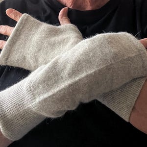 Może przedstawiać: Para jasnoszarych rękawiczek bez palców. Rękawiczki wykonane są z miękkiego, puszystego materiału i mają prążkowane mankiety. Rękawiczki mają zakrywać dłoń i nadgarstek, pozostawiając palce odsłonięte.