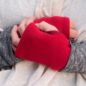 Może przedstawiać: Para czerwonych rękawic bez palców z czarnym wykończeniem. Rękawice są złożone na pół i trzymane w dłoniach osoby w szarym swetrze.