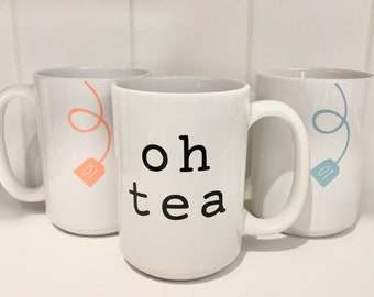 May include: Three white ceramic mugs with black text and a tea bag design. The mug in the center says "oh tea" and has a pink tea bag. The mug on the left has a pink tea bag and the mug on the right has a blue tea bag.