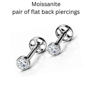 May include: A pair of silver-toned flat back piercings featuring round, clear Moissanite stones set in a bezel setting. The text Moissanite pair of flat back piercings is displayed above the jewelry, which has circular cutouts on the backings.