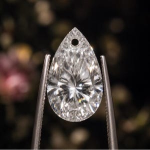 Peut inclure: Un pendentif en pierre précieuse incolore et transparent en forme de poire, doté d'une surface à facettes qui reflète la lumière. La pierre présente un petit trou circulaire percé près de la pointe supérieure, tenue par des pinces métalliques.