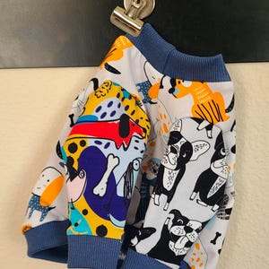 May include: Two pairs of dog-themed baby pants with blue cuffs and waistbands. One pair features colorful cartoon dogs, bones, and abstract shapes. The other pair has a pattern of black and white French Bulldogs.