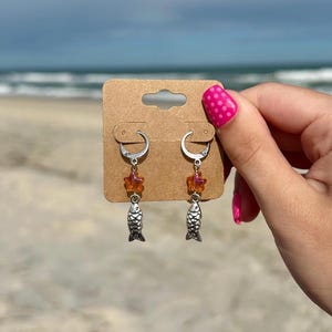 May include: A pair of silver hoop earrings with dangling fish charms. Each earring features a small orange star-shaped bead above the fish. The earrings are displayed on a brown card, with a beach and ocean background.