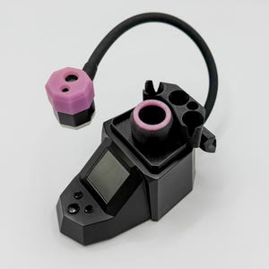 Puede incluir: Un dispositivo electrónico negro con una pantalla y botones, conectado a un tubo negro curvado y un componente rosa y negro. El dispositivo tiene un diseño geométrico y una abertura circular con interior rosa.