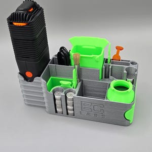 May include: A gray and green organizer with compartments for a black vaporizer, a brush, a small container, and a set of metal screens. The organizer has the text "BC Labs" printed on the front.