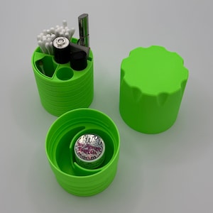 Może przedstawiać: Zielony plastikowy pojemnik z pokrywką, zawierający różne przedmioty, w tym patyczki kosmetyczne, małą metalową rurkę i baterię. Pojemnik jest podzielony na sekcje. Pokrywka jest również zielona i ma fakturowaną powierzchnię.