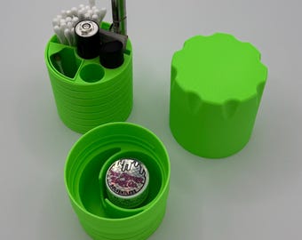 Może przedstawiać: Zielony plastikowy pojemnik z pokrywką, zawierający różne przedmioty, w tym patyczki kosmetyczne, małą metalową rurkę i baterię. Pojemnik jest podzielony na sekcje. Pokrywka jest również zielona i ma fakturowaną powierzchnię.