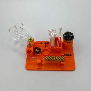 May include: An orange silicone dab station with various compartments and a glass rig. The station includes a section for cotton swabs, a small container, and a holder for a glass tool. The text "EG LABS" is printed on the front.