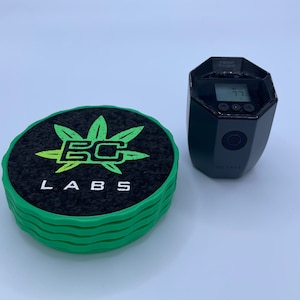 Puede incluir: Un conjunto de cuatro posavasos verdes y negros con el logotipo "BC Labs" y un diseño de hoja de marihuana. Un temporizador digital negro con un botón azul y la palabra "Octave" está al lado de los posavasos.