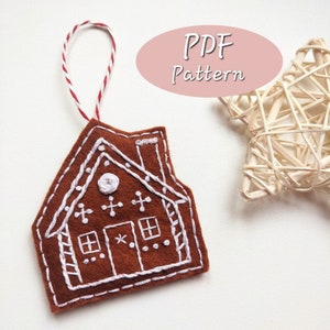 Puede incluir: Un adorno de casa de jengibre de fieltro marrón con detalles bordados en blanco, incluyendo una chimenea, ventanas y un diseño de bastón de caramelo. El adorno tiene un lazo de cuerda rojo y blanco para colgar. El texto "PDF Pattern" es visible en el fondo.
