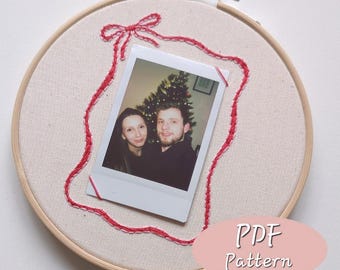 May include: An embroidery hoop with a cream-colored fabric background. A red embroidered frame surrounds a Polaroid photo of a couple. The photo is secured with red thread. The text "PDF Pattern" is in a pink oval.