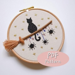 Peut inclure: Art de broderie en forme de cercle représentant un chat noir assis sur une branche avec un balai et des araignées suspendues. Le fond est un tissu beige avec des étoiles dorées. Le texte "PDF Pattern" est sur un ovale rose.