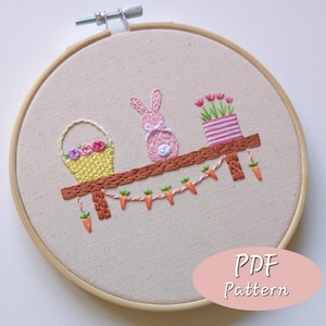 Puede incluir: Bordado en bastidor con temática de Pascua con un conejito rosa, una cesta de flores y tulipanes en maceta sobre una estantería de madera. Una guirnalda de zanahorias cuelga debajo. El texto "PDF Pattern" se muestra en un óvalo rosa.