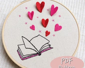 May include: A white embroidery hoop with a black line drawing of an open book and pink and red felt hearts floating above it. The text "PDF Pattern" is in the lower right corner of the hoop.