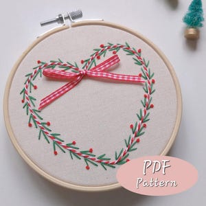 May include: Embroidered heart-shaped wreath on a cream-colored fabric, framed in a wooden hoop. The wreath features green and red stitching with a red and white checkered ribbon bow. The text "PDF Pattern" is in a pink oval.