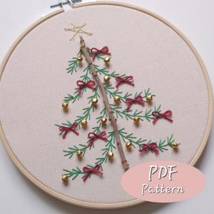 Könnte beinhalten: Gesticktes Weihnachtsbaum-Design auf cremefarbenem Stoff, gespannt in einem Holzreifen. Der Baum hat grüne, gestickte Zweige, rote Schleifen, goldene Ornamente und einen goldenen Stern. Der Text "PDF Pattern" befindet sich in einem rosa Oval.