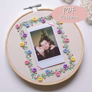 May include: A round embroidery hoop with a cream-colored fabric background. A photo is centered, surrounded by a floral embroidery design in pink, yellow, blue, and purple. The text "PDF Pattern" is in a pink oval.
