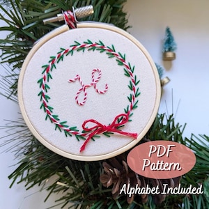 以下が含まれることがあります： 白の刺繍枠に、赤と緑のリースのデザインと中央に「H」の文字が刺繍されています。枠の周りは緑のガーランドと松ぼっくりで飾られています。枠の下には「PDF Pattern Alphabet Included」の文字が見えます。