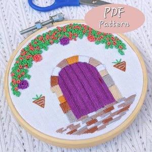 May include: Embroidered hoop art featuring a purple door with a brick archway and a green vine with pink flowers above. The hoop is made of wood and has a white fabric background.