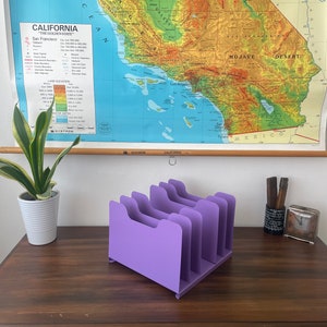 May include: A lavender desk organizer with six vertical slots. The organizer is made of a solid material and sits on a dark wooden desk. A map of California hangs on the wall behind the desk.