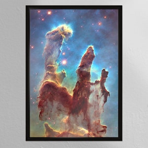 May include: A framed print of a colorful nebula with tall, thin pillars of gas and dust. The nebula is a vibrant mix of red, orange, blue, and white, with a bright star shining in the center.