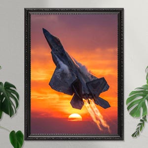 May include: Framed print featuring a grey F-22 fighter jet ascending against a vivid sunset. The jet is angled upwards, with vapour trails trailing from its engines. The sun is a bright orange orb near the horizon. The print is in a black frame.