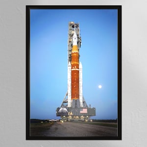 May include: Framed print of a large rocket on a launchpad, set against a blue sky with a full moon. The rocket is primarily orange and white, with the American flag visible. The print is from MINIBOT Print Shop.