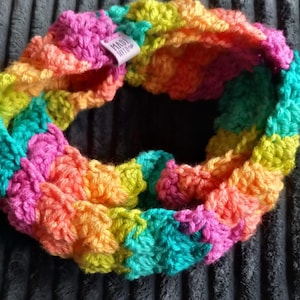 Könnte beinhalten: Ein farbenfroher, gehäkelter Loop-Schal in leuchtenden Farben wie Pink, Gelb, Orange und Türkis. Der Schal ist handgefertigt, wie ein kleines Etikett mit der Aufschrift "MADE WITH" zeigt. Der Schal ist eine Endlosschleife.