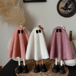 May include: Three handmade ghost decorations made from fabric. The ghosts are pink, white, and pink. Each ghost has black eyes and a small flower on top of its head. The ghosts are standing on a woven rug.