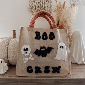 Könnte beinhalten: Eine braune Jute-Tragetasche mit einem schwarzen Filz-Design "BOO CREW". Die Tasche hat einen weißen Filz-Geist, eine schwarze Filz-Fledermaus mit gelben Augen und einen weißen Filz-Schädel mit Kreuzknochen.