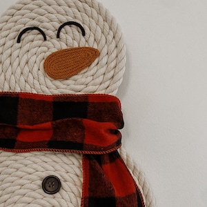 May include: A white rope snowman with a red and black plaid scarf and a brown felt nose. The snowman has black eyes and a smiling mouth.