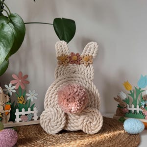 Pode incluir: Um coelho de corda branca com uma cauda de pompom rosa e uma coroa de flores de fio rosa e amarelo. O coelho está sentado em um tapete tecido marrom.