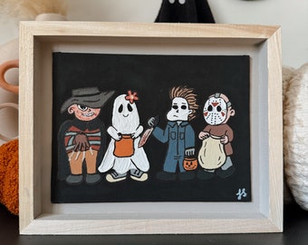 May include: A framed Halloween-themed painting featuring four cartoon characters: a cowboy, a ghost, Michael Myers, and Jason Voorhees. The characters are depicted against a black background, each holding a prop associated with their character. The frame is made of light-colored wood.