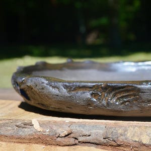 May include: A handmade, dark brown ceramic bowl with a rough, textured surface. The bowl has an organic, irregular shape with a slightly raised rim. The bowl is approximately 12 inches in diameter.
