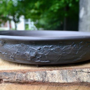 May include: A shallow, circular dark gray ceramic bonsai pot with a matte finish. The exterior wall features a textured, rugged, and uneven surface pattern that contrasts with the smooth, flat interior and the clean, rounded rim of the vessel.