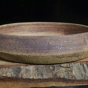 May include: A shallow, circular ceramic bowl with a wide, angled rim. The exterior features a textured, speckled finish in earthy tones of tan, brown, and dark blue, with a matte appearance and visible clay grain throughout the surface.