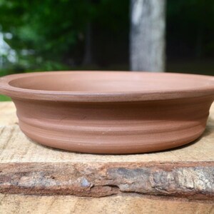May include: A shallow, circular terracotta bowl with a smooth, matte finish and a wide, flat rim. The exterior wall of the bowl features subtle, horizontal ridged detailing that encircles the base, providing a simple, uniform texture.