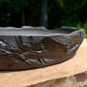 May include: A shallow, circular dark gray ceramic tray with a textured, uneven outer rim. The surface of the clay features deep, carved grooves and ridges that create a rugged, organic appearance around the entire perimeter of the vessel.