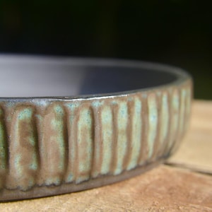 May include: A shallow ceramic bowl with a smooth, dark interior and a textured exterior featuring vertical ridges. The outer surface displays a mottled finish with shades of brown and a weathered, oxidized teal green glaze in the grooves.