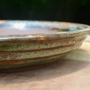 May include: A shallow, circular ceramic dish featuring a textured, porous rim with a mottled glaze in shades of earthy brown, muted green, and hints of blue. The exterior side displays horizontal ridges and a rough, pitted surface finish.