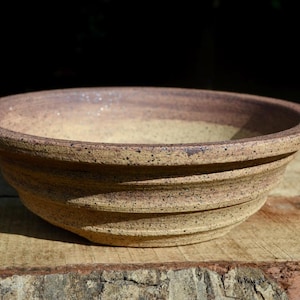 May include: A shallow, round ceramic bowl featuring a speckled beige and brown glaze. The exterior surface displays horizontal ridges or grooves circling the body, while the interior has a smooth, concave shape with a wide, flat rim.