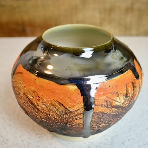 May include: A handcrafted ceramic vase with a wide, rounded body and a narrow opening. The vase features a textured orange and brown exterior with dark blue and black accents. The interior is a light green color.
