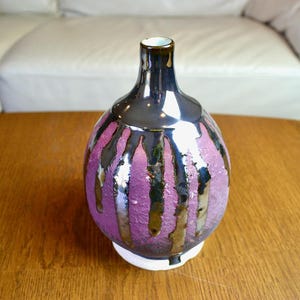 May include: A decorative ceramic vase with a bulbous body and a narrow neck. The vase features a glossy black glaze with vertical drips of gold and purple. The base is white, and the vase sits on a wooden surface.