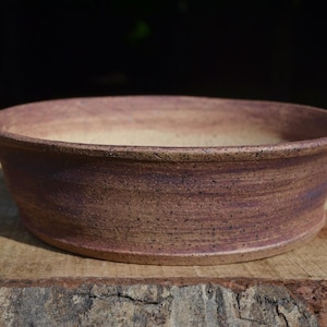 May include: A round, shallow ceramic bowl with a textured, mottled exterior in shades of dusty rose and brown. The interior of the bowl features a smooth, light beige finish with subtle speckling throughout the clay surface.
