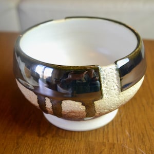 May include: A ceramic bowl with a white interior and a metallic, dark brown exterior. The bowl has a textured, light-colored band and a white base. The bowl is on a wooden surface.