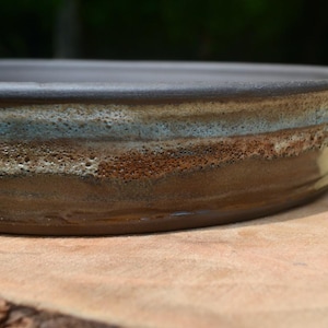 May include: A shallow, round ceramic tray featuring a dark brown base color with a textured, layered glaze. The rim displays horizontal bands of mottled light blue, cream, and earthy brown tones, creating a rugged, uneven surface finish.