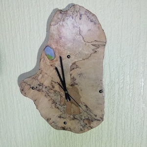 May include: A wooden clock with a unique, irregular shape. The clock face is made of natural wood with a light brown colour and dark brown veins. The clock hands are black and the clock has a small painted landscape scene on the top.
