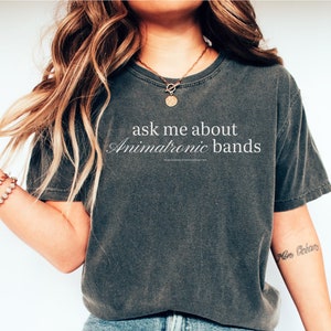 May include: A woman wearing a dark gray t-shirt with the text "ask me about animatronic bands" printed on it. She is wearing blue jeans and white sneakers.